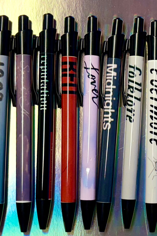 Fourteen vibrant, refillable Eras Pens by Kreative Kreationz are neatly lined up on a metallic surface. Each pen, filled with black gel ink and adorned with album titles from 