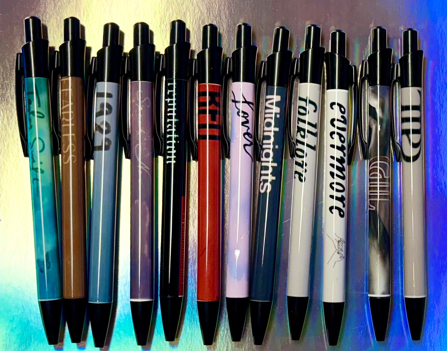 Fourteen vibrant, refillable Eras Pens by Kreative Kreationz are neatly lined up on a metallic surface. Each pen, filled with black gel ink and adorned with album titles from "Taylor Swift" to "Midnights," celebrates Taylor Swift's Eras, exuding charm and style in black and white script.