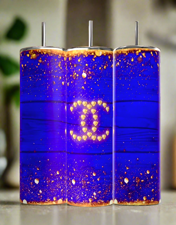 The Kreative Kreationz Chanel 20oz Skinny Tumbler in royal blue features a bold blue and gold glitter design with a gold Chanel logo, complemented by metal straws—luxurious, elegant drinkware perfect for any occasion.