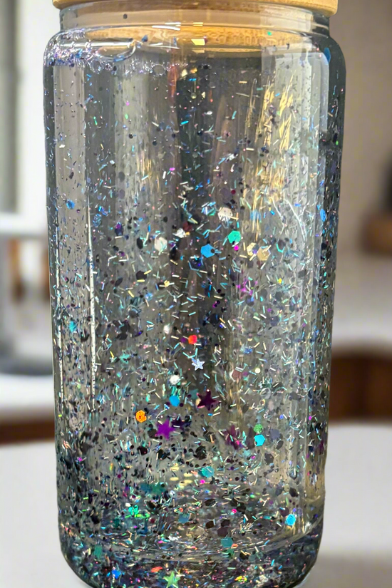 The Snowglobe Tumbler by Kreative Kreationz is a clear glass jar with a bamboo lid, filled with water and floating multicolored glitter, including silver stars—a charming handmade accent for any space.