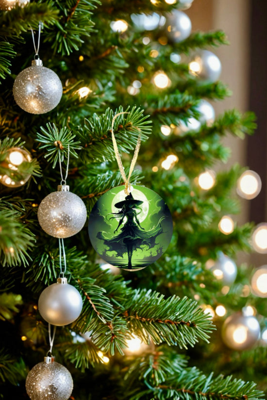A close-up of a decorated Christmas tree with silver baubles and white lights features the special Wicked Witch Christmas Tree Ornament by Kreative Kreationz. This distinctive ornament showcases a sublimated design with the silhouette of a person set against a green, mystical background, adding an enchanting touch to your holiday decor.