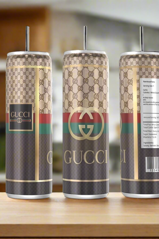 Three Gucci Silver Stainless Steel Inspired 20oz Tumblers by Kreative Kreationz, each with a metal straw and eco-friendly reusable design, rest on wood; one features a faux nutrition label against a blurred kitchen backdrop.