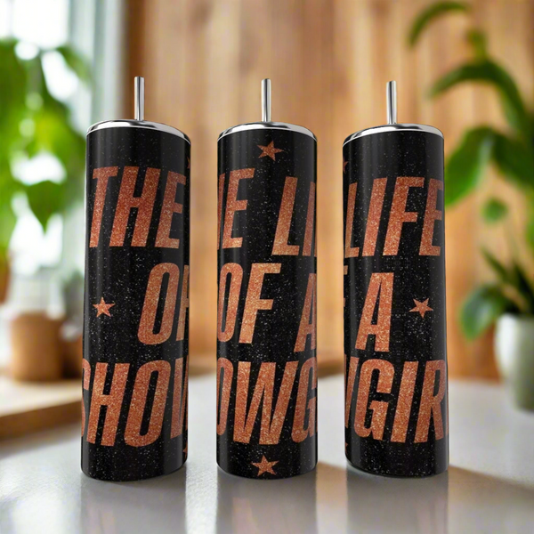 Three black glitter stainless steel "The life of a Showgirl Taylor Swift 20oz Tumblers" by Kreative Kreationz, each with metallic lid and straw, are on a table. Copper text stands out; plants and wood paneling blur in the background.