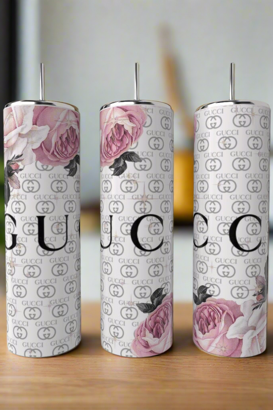 Three tall Kreative Kreationz Gucci Inspired 20oz Tumblers, featuring pink rose designs and repeating logo patterns, are displayed on a wooden surface in a blurred indoor setting, highlighting stylish luxury drinkware.