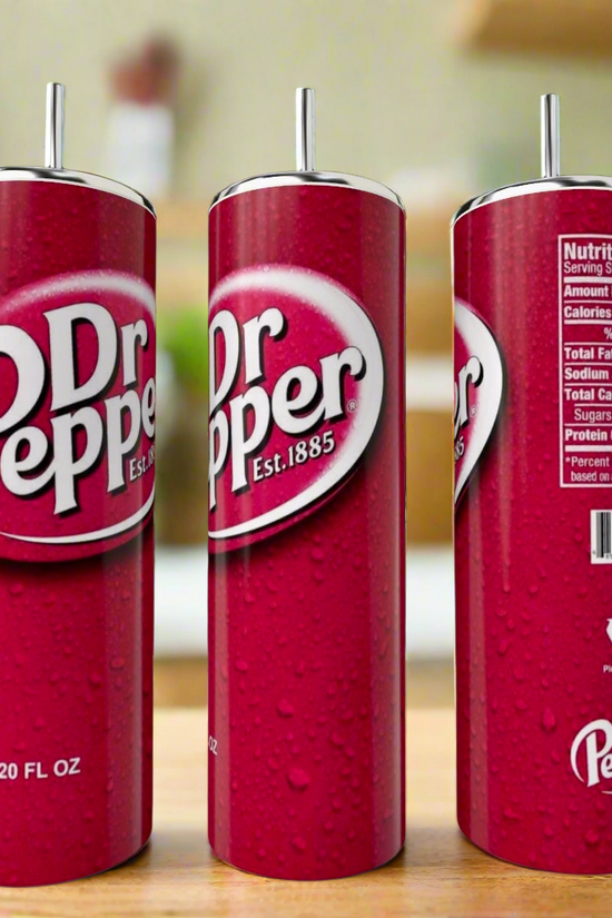 A Dr Pepper Stainless Steel Tumbler by Kreative Kreationz sits on a kitchen counter with three tall Dr Pepper cans nearby, showing the logo, nutrition facts, and recycling symbol against a blurred kitchen background.