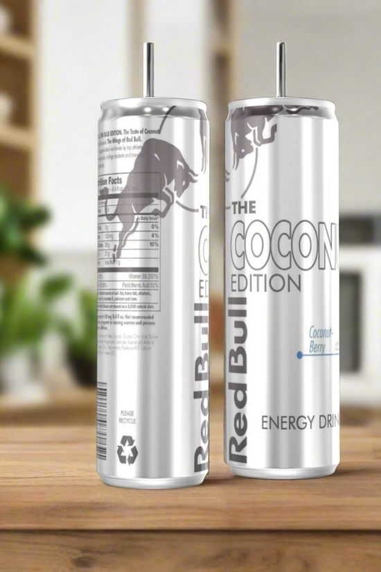 Two Coconut Red Bull Stainless Steel 20oz Tumblers by Kreative Kreationz sit on a wooden surface, one showing the product name and the other with nutritional info, both set against a softly blurred background.