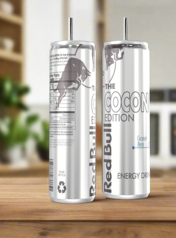 Two Coconut Red Bull Stainless Steel 20oz Tumblers by Kreative Kreationz sit on a wooden surface, one showing the product name and the other with nutritional info, both set against a softly blurred background.
