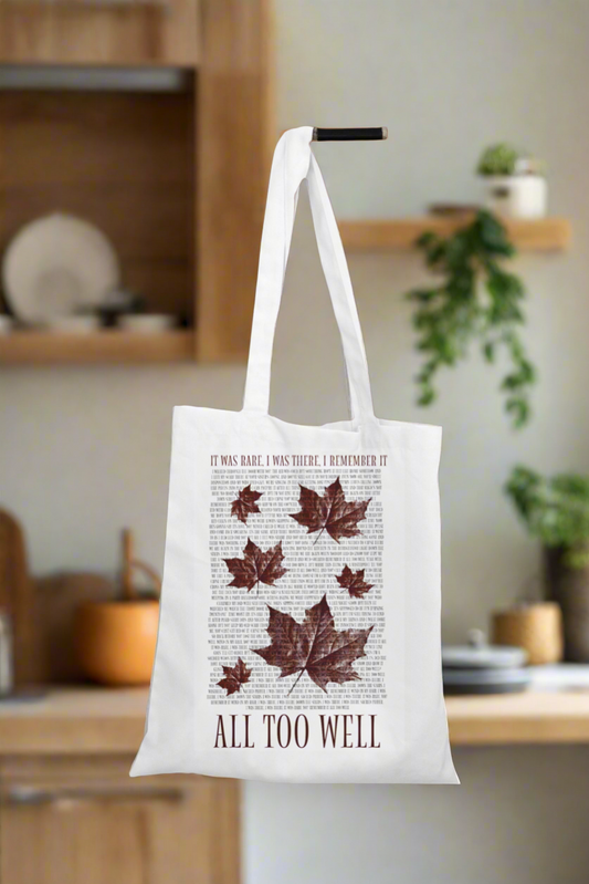 In a kitchen setting, a white "All Too Well Tote Bag with lyrics" by Kreative Kreationz hangs, featuring text and illustrations of autumn leaves. The bag reads, "It was rare, I was there, I remember it," with "All Too Well" by Taylor Swift at the bottom; kitchenware and a potted plant complete the scene.
