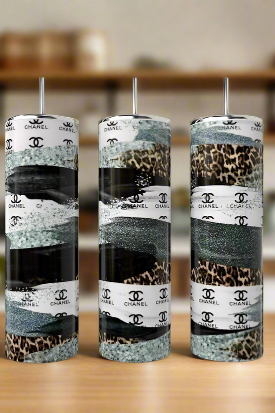 On a wooden surface sit three tall Kreative Kreationz 20oz stainless steel tumblers with metallic lids, available in glitter, leopard print, and Chanel stripe designs. In the background, blurred shelves emphasize the tumblers' sophisticated elegance.