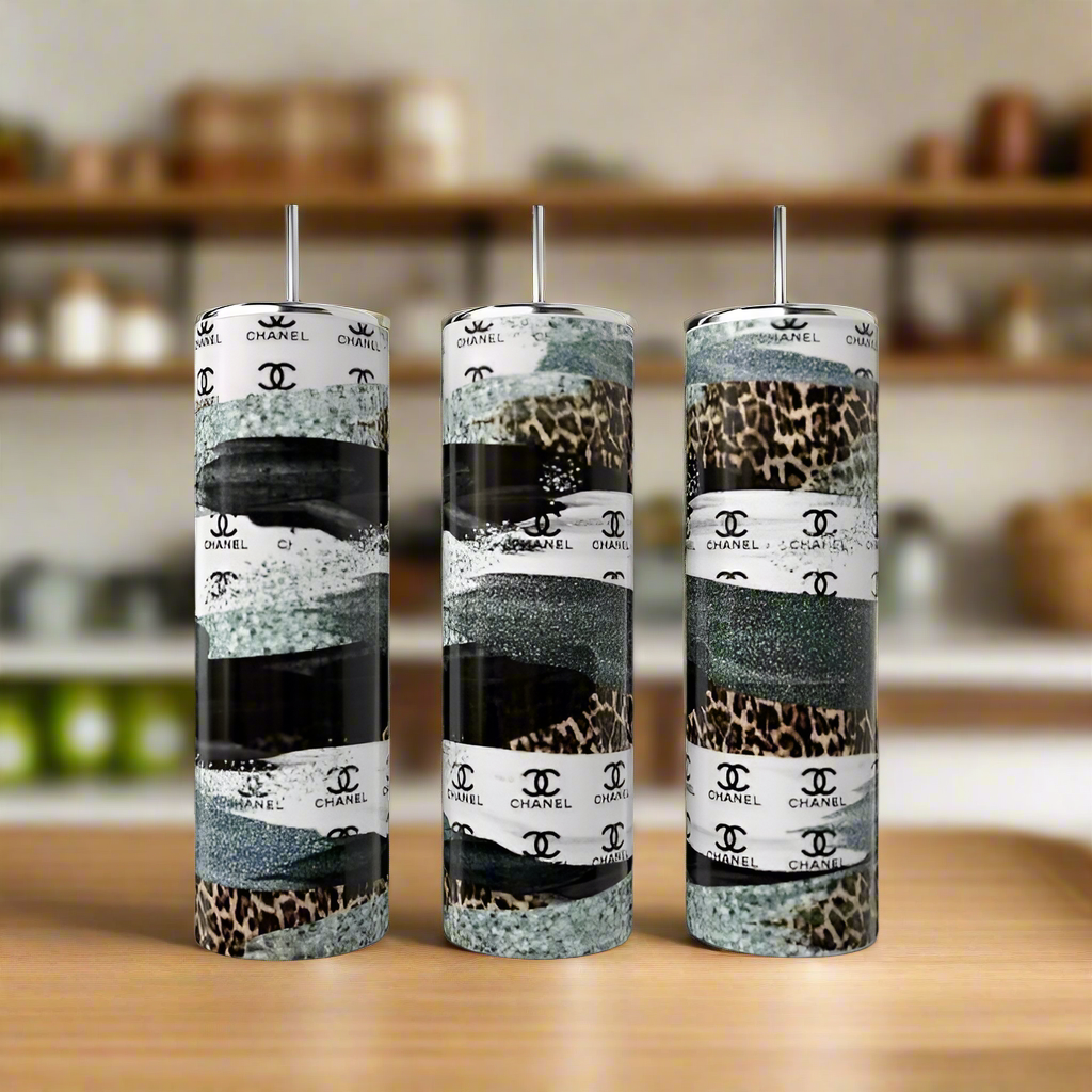 On a wooden surface sit three tall Kreative Kreationz 20oz stainless steel tumblers with metallic lids, available in glitter, leopard print, and Chanel stripe designs. In the background, blurred shelves emphasize the tumblers' sophisticated elegance.