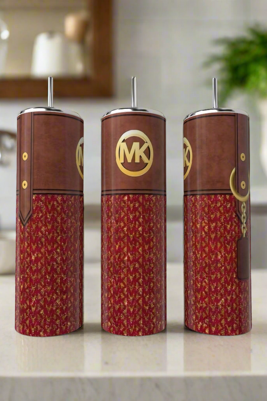 Three MK 20oz Tumblers in brown and red by Kreative Kreationz with gold logos and metallic spouts are on a kitchen counter, resembling Michael Kors products. A faucet, shelf, and green plant appear blurred in the background.