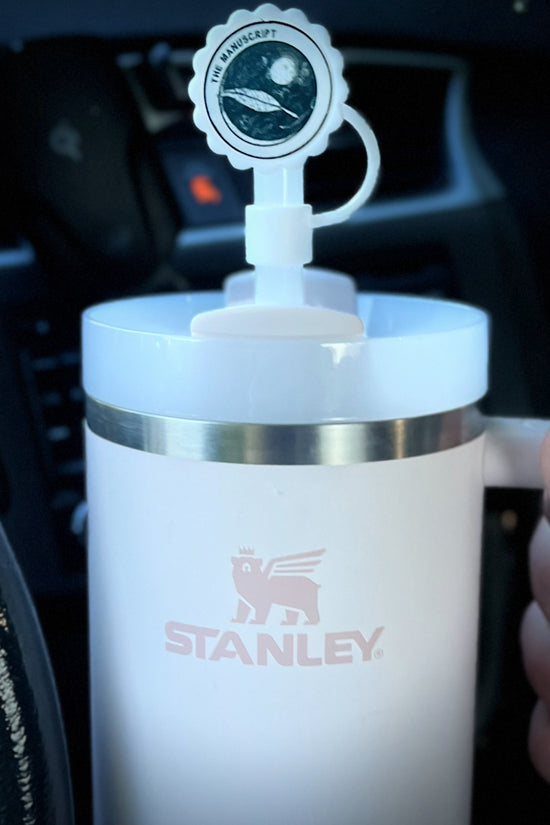 Stanley cup with a white straw cap that’s circular on the circular cap it says “the manuscript”  with a feather in dark writing 