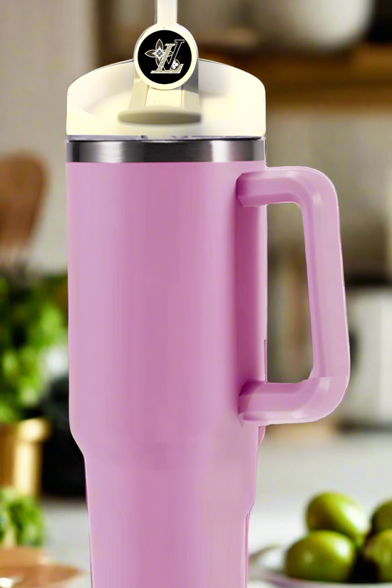 A pink insulated tumbler with a lid and Stanley tumbler accessory adorns the kitchen counter, its straw showcasing the Kreative Kreationz LV Straw Topper. In the background, blurred plants and small green fruits bring a natural touch to the scene.