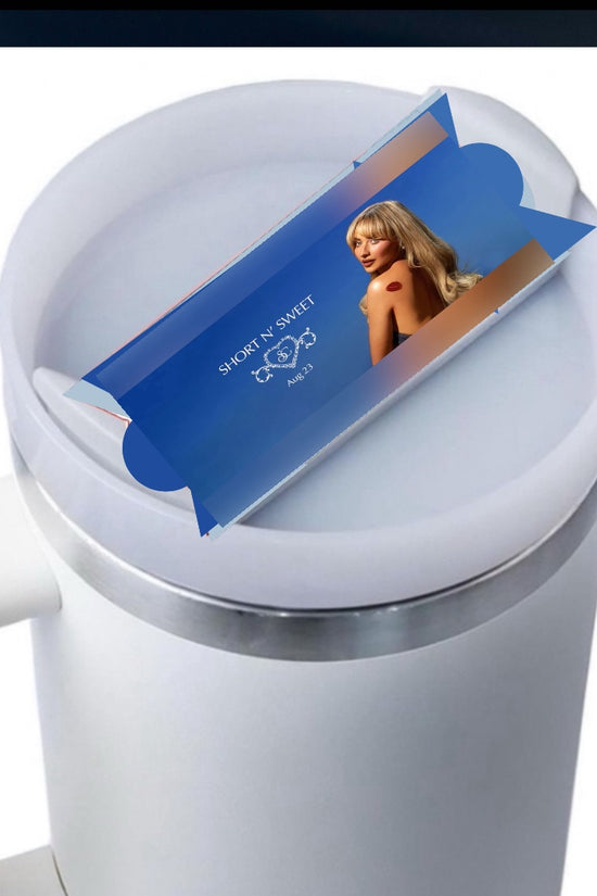 A white mug with a light blue button feature, covered by a lid. On the lid sits a blue, rectangular packaging featuring an image of a woman with blonde hair in a black outfit. The text on the packaging reads 