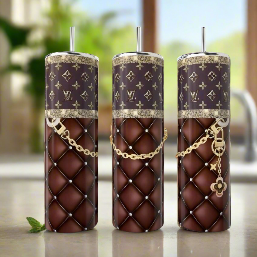 Three LV Handbag Inspired 20oz Tumblers by Kreative Kreationz, featuring quilted brown and gold chain designs with patterned tops, are elegantly displayed on a kitchen counter with mint leaves nearby.