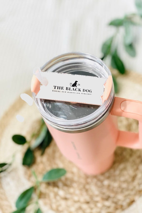 A pink tumbler with a TTPD Black Dog Stanley Lid Plate/Topper from Kreative Kreationz sits on a woven mat. On top of the lid, there is a white sticker with black text reading 