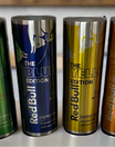 Four tall cans of Red Bull editions in green, blue, yellow, and amber colors are lined up on a table. Beside them is a sleek RB Energy Silver Tumbler by Kreative Kreationz, with 20 oz capacity—ideal for keeping your favorite edition ice-cold and ready to energize.