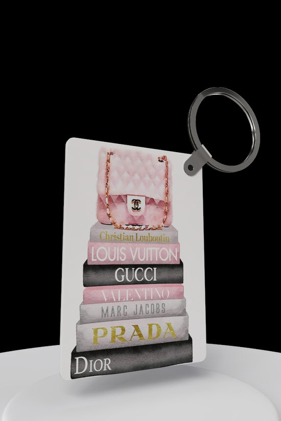 The Designer Inspired Keychain by Kreative Kreationz features a pink quilted purse atop stacked luxury brand books against a black background—a stylish accessory and perfect gift for fashion lovers!.