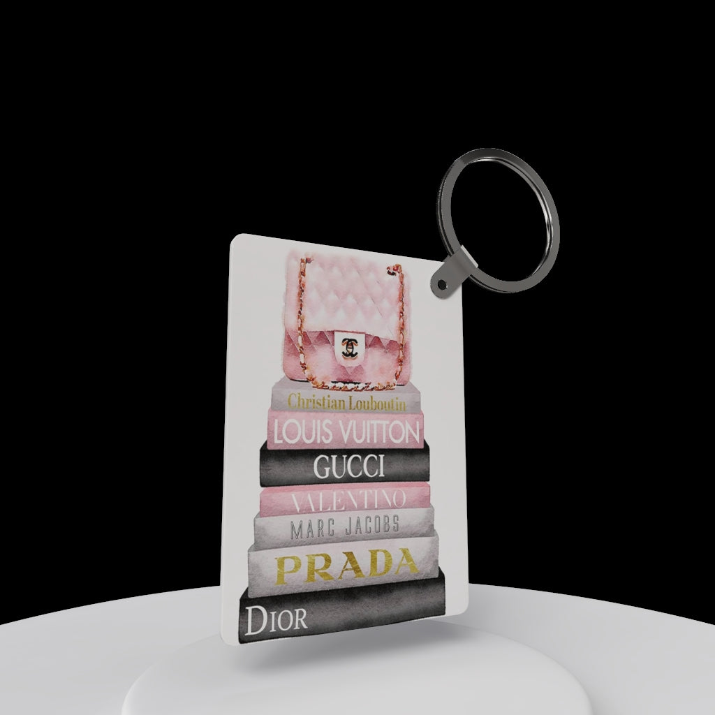 The Designer Inspired Keychain by Kreative Kreationz features a pink quilted purse atop stacked luxury brand books against a black background—a stylish accessory and perfect gift for fashion lovers!.