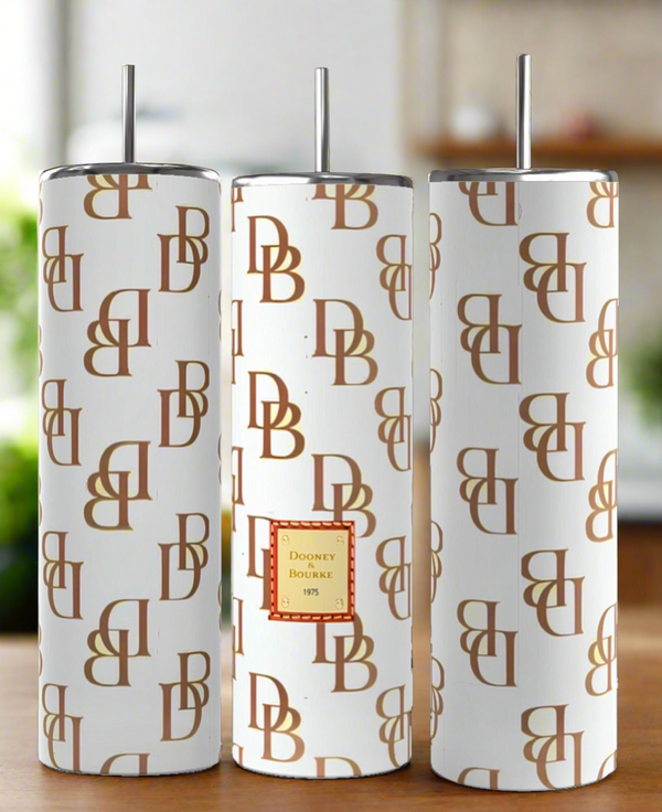 Three D&B -White | Brown 20oz Tumblers by Kreative Kreationz with brown monogram patterns and silver straws are shown. The center tumbler boasts a red and gold "Dooney & Bourke 1975" label. Background is softly blurred.