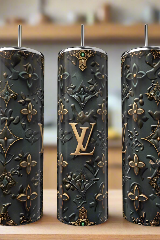 The LV 20oz Tumbler by Kreative Kreationz features a tall, cylindrical stainless steel design with straws and an embossed gold and black pattern, adding a touch of luxury to your drinkware collection. Displayed on wood with a blurred kitchen background.