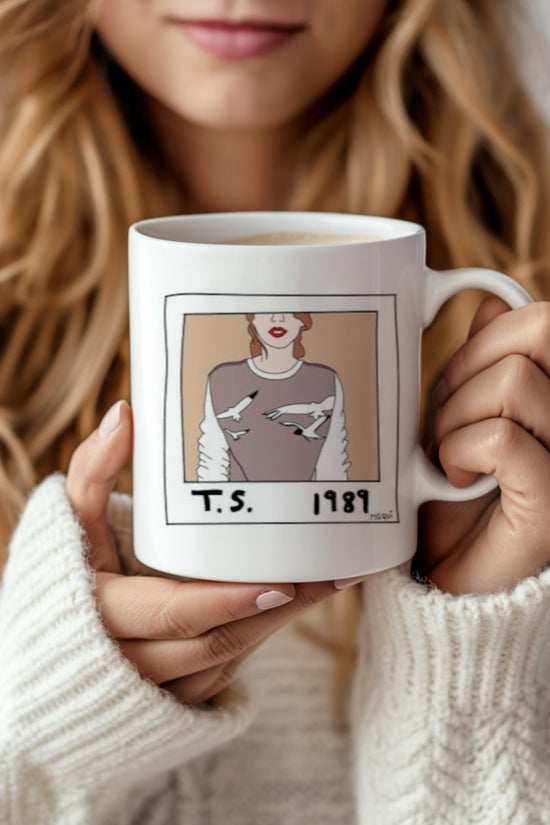 A woman with wavy blonde hair in a white sweater holds the 1989 Polaroid Ceramic Mug by Kreative Kreationz, featuring an illustrated woman in a bird-patterned sweater with 