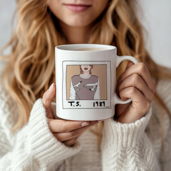 A woman with wavy blonde hair in a white sweater holds the 1989 Polaroid Ceramic Mug by Kreative Kreationz, featuring an illustrated woman in a bird-patterned sweater with "T.S. 1989"—a perfect gift for any Swiftie.