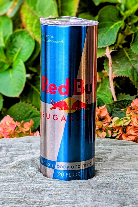A 20 oz RB Energy- Silver Stainless Steel Tumbler by Kreative Kreationz, showcasing its signature logo and design, sits on a cloth surface outdoors amidst green leaves and pink flowers, creating a serene garden atmosphere.