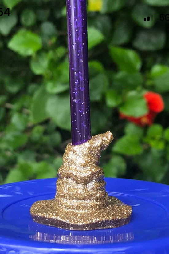 A Harry Potter Wizard | Witch Hat straw Topper by Kreative Kreationz, featuring a purple sparkly pencil in gold glittery clay on a blue bottle cap, stands against blurred greenery and red flowers, capturing the whimsical charm of Harry Potter accessories.
