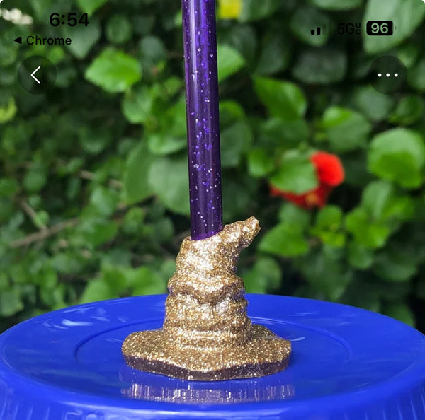 A Harry Potter Wizard | Witch Hat straw Topper by Kreative Kreationz, featuring a purple sparkly pencil in gold glittery clay on a blue bottle cap, stands against blurred greenery and red flowers, capturing the whimsical charm of Harry Potter accessories.