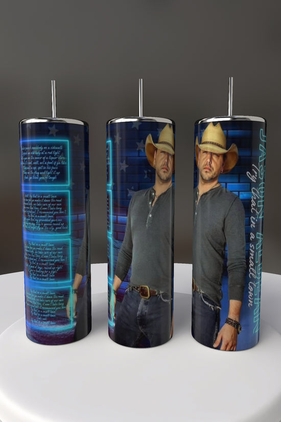 Set against a dark background, the Jason Aldean 20oz Tumblers by Kreative Kreationz display a man in a cowboy hat, gray shirt, and jeans with blue digital text and graphics. These eco-friendly stainless steel tumblers feature an exclusive design.