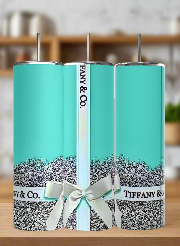 Three 20oz Tiffany Tumblers by Kreative Kreationz feature blue designs, black and white glitter accents, silver stainless steel lids, straws, and "Tiffany & Co." print. Center tumbler has a white ribbon. Blurred kitchen background.
