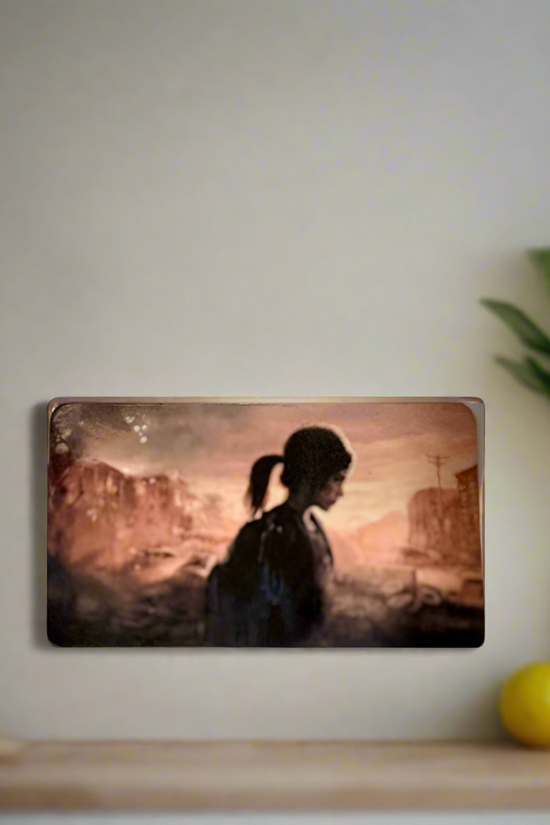 A portrait of a ponytailed figure in profile hangs on a beige wall. Nearby, gaming collectibles stand out—a Kreative Kreationz The Last Of Us Fridge Magnet sits on the shelf beside a green plant and yellow ball against a desolate landscape backdrop.