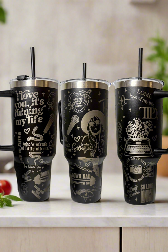Three large black Kreative Kreationz Taylor Swift TTPD 40oz Engraved Tumblers with handles sit on a kitchen counter. Each stainless steel mug has white Taylor Swift-themed designs. A blurred kitchen scene with tomatoes is in the background.