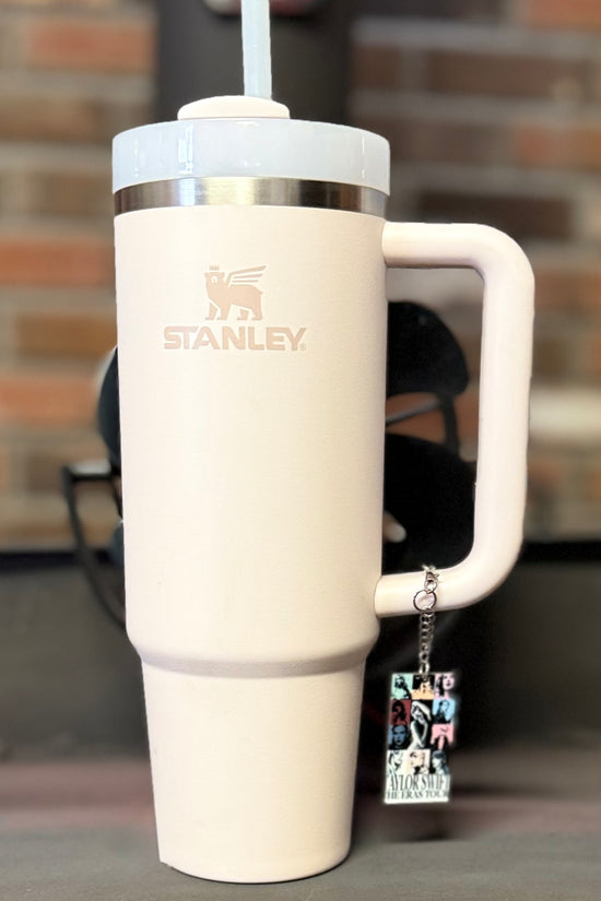 A large, light pink Stanley tumbler with a straw stands on a surface. Hanging from the handle is the ERAS Charm Dangle cup accessory by Kreative Kreationz, featuring black and white images and text. A blurred brick wall forms the background.