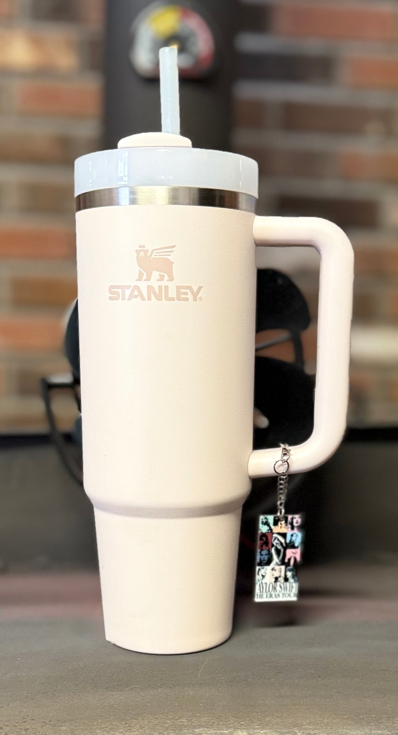 A large, light pink Stanley tumbler with a straw stands on a surface. Hanging from the handle is the ERAS Charm Dangle cup accessory by Kreative Kreationz, featuring black and white images and text. A blurred brick wall forms the background.