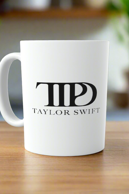 A white TTPD All’s Fair in Love and Poetry | Who’s Afraid of Little Old Me ceramic mug by Kreative Kreationz sits on wood, featuring a bold black logo—a perfect Swiftie gift. The softly blurred background adds charm.