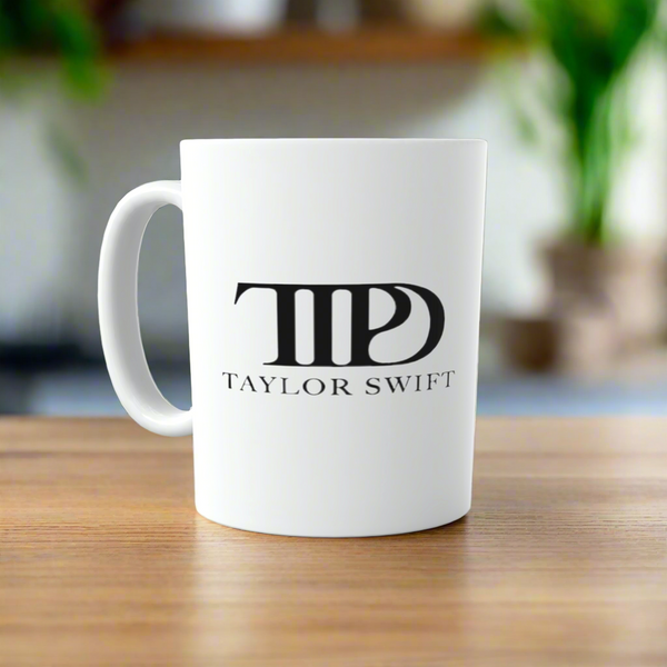 A white TTPD All’s Fair in Love and Poetry | Who’s Afraid of Little Old Me ceramic mug by Kreative Kreationz sits on wood, featuring a bold black logo—a perfect Swiftie gift. The softly blurred background adds charm.