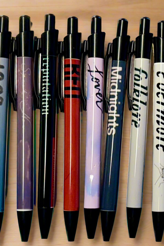 Fourteen Eras Pens by Kreative Kreationz are lined up on a wooden surface. Each refillable black gel pen displays designs and text inspired by Taylor Swift album titles like 