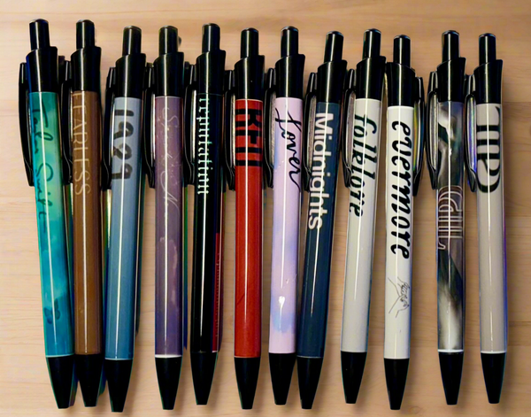 Fourteen Eras Pens by Kreative Kreationz are lined up on a wooden surface. Each refillable black gel pen displays designs and text inspired by Taylor Swift album titles like "Red," "Lover," "Midnights," "Folklore," and "Evermore.