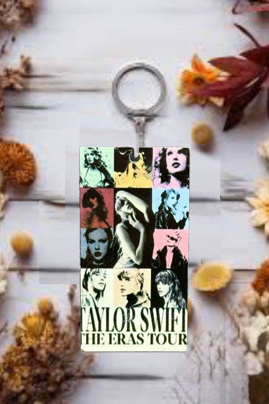 The ERAS Keychain by Kreative Kreationz displays vibrant portraits of Taylor Swift from various eras, with “Taylor Swift The Eras Tour” text, resting on a light wooden surface accented by dried flowers.