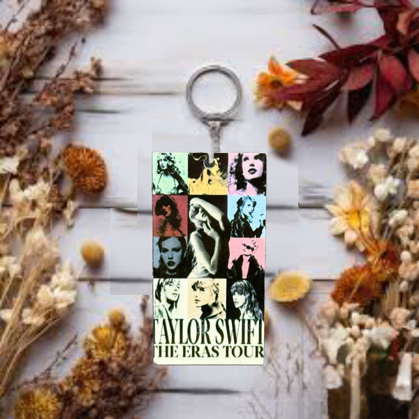The ERAS Keychain by Kreative Kreationz displays vibrant portraits of Taylor Swift from various eras, with “Taylor Swift The Eras Tour” text, resting on a light wooden surface accented by dried flowers.