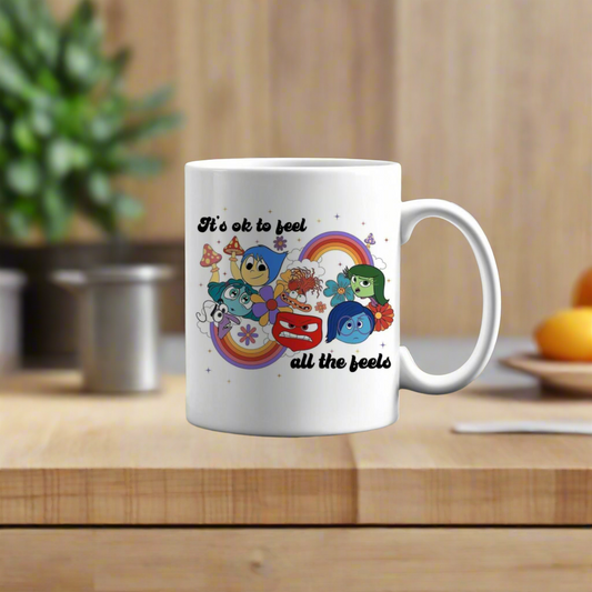 The Feels Coffee Ceramic Mug by Kreative Kreationz features colorful cartoon emotions, rainbows, stars, and “It’s ok to feel all the feels”—a unique gift for any occasion. Blurred plants and fruit set a cozy kitchen vibe in the background.
