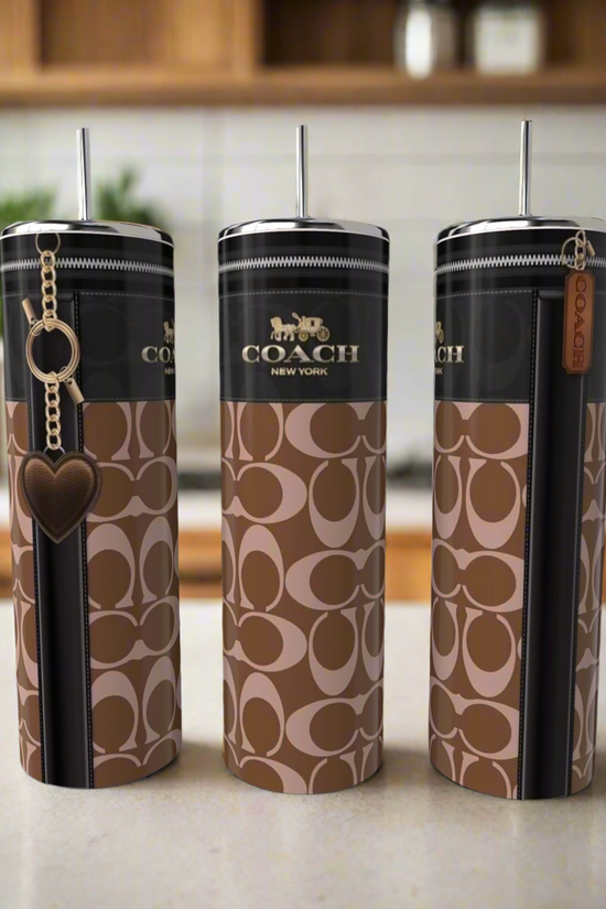 Three stylish, eco-friendly C Buckle 20oz Skinny Tumblers by Kreative Kreationz, featuring a brown and black pattern and made of high-quality stainless steel, grace the kitchen counter. Each tumbler sports a unique charm: a heart, zipper pull, and leather tag—a perfect blend of luxury-inspired design.