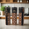 Three stylish, eco-friendly C Buckle 20oz Skinny Tumblers by Kreative Kreationz, featuring a brown and black pattern and made of high-quality stainless steel, grace the kitchen counter. Each tumbler sports a unique charm: a heart, zipper pull, and leather tag—a perfect blend of luxury-inspired design.