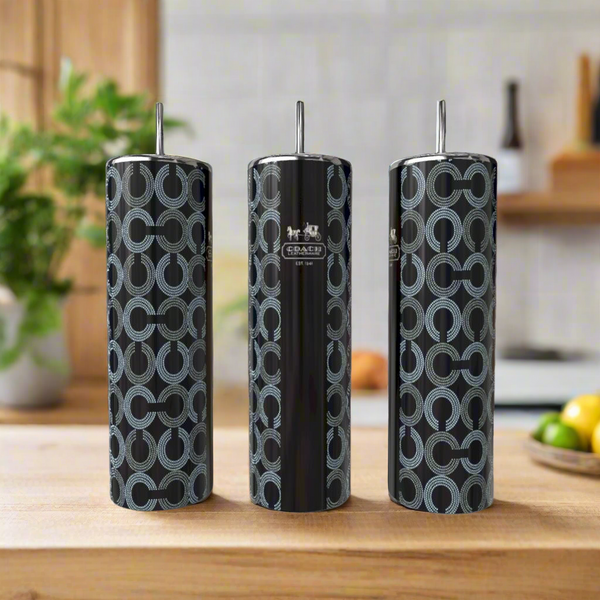 Three tall, cylindrical Coach Inspired 20oz Tumblers by Kreative Kreationz with silver lids sit on a wooden countertop. Their black base has a blue circular pattern and logo detail. A blurred kitchen is visible in the background.