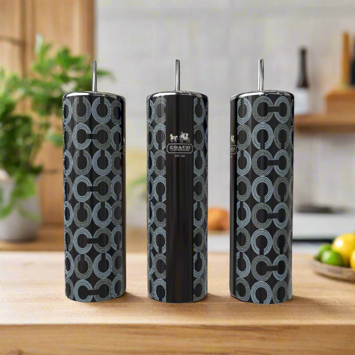 Three tall, cylindrical Coach Inspired 20oz Tumblers by Kreative Kreationz with silver lids sit on a wooden countertop. Their black base has a blue circular pattern and logo detail. A blurred kitchen is visible in the background.