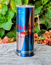 A Sugar Free Red Bull 20oz Tumbler by Kreative Kreationz stands upright on a gray cloth outdoors, with green leaves and pink hydrangea flowers in the background.