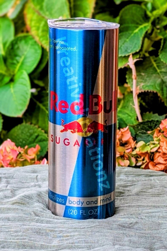 A Sugar Free Red Bull 20oz Tumbler by Kreative Kreationz stands upright on a gray cloth outdoors, with green leaves and pink hydrangea flowers in the background.