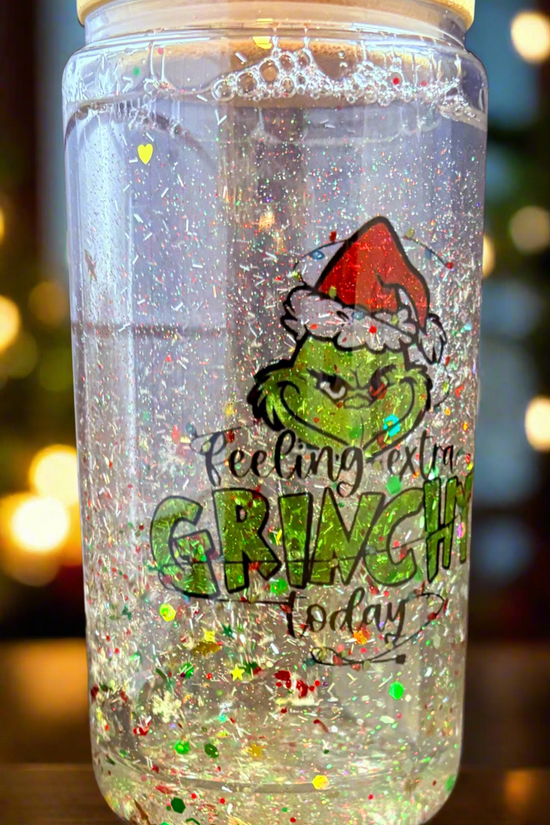 The Feeling Extra Grinchy Today Snowglobe Tumbler from Kreative Kreationz features a festive Grinch illustration with a Santa hat, filled with clear liquid and glitter. It's the perfect Christmas gift, enhanced by softly blurred warm lights.